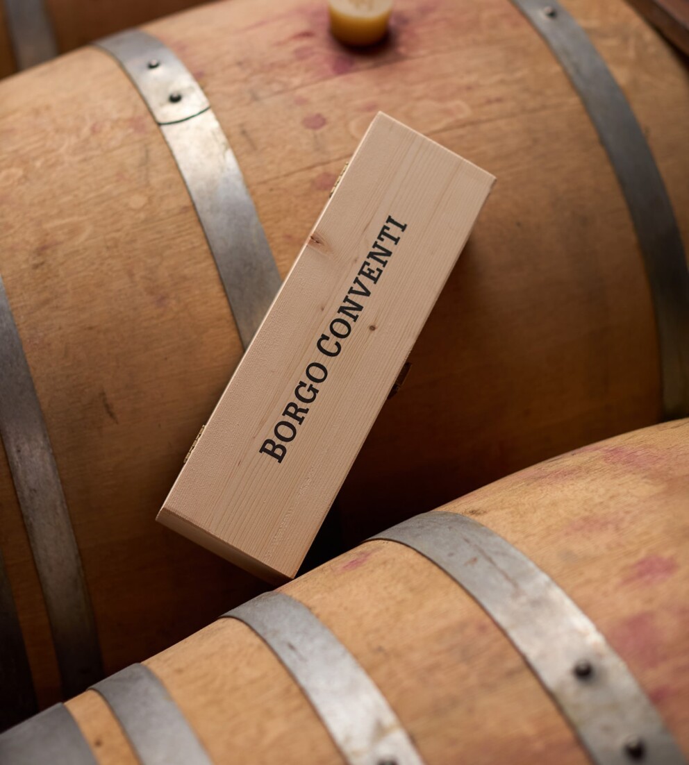 Cofanetto chiuso in legno di Borgo Conventi contenete una bottiglia di vino, appoggiato sopra a delle botti di legno